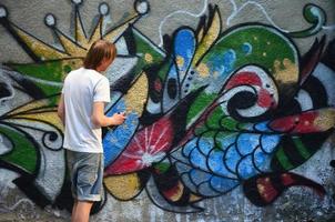 foto van een jong vent in denim shorts en een wit shirt. de vent trekt Aan de graffiti muur een tekening met aërosol verven van divers kleuren. de concept van hooliganisme en schade naar eigendom