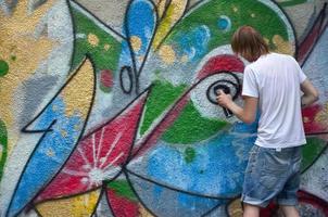 foto van een jong vent in denim shorts en een wit shirt. de vent trekt Aan de graffiti muur een tekening met aërosol verven van divers kleuren. de concept van hooliganisme en schade naar eigendom