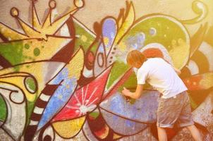 foto van een jong vent in denim shorts en een wit shirt. de vent trekt Aan de graffiti muur een tekening met aërosol verven van divers kleuren. de concept van hooliganisme en schade naar eigendom