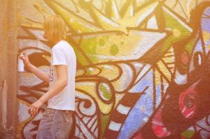foto van een jong vent in denim shorts en een wit shirt. de vent trekt Aan de graffiti muur een tekening met aërosol verven van divers kleuren. de concept van hooliganisme en schade naar eigendom