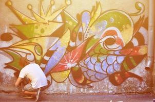 foto van een jong vent in denim shorts en een wit shirt. de vent trekt Aan de graffiti muur een tekening met aërosol verven van divers kleuren. de concept van hooliganisme en schade naar eigendom