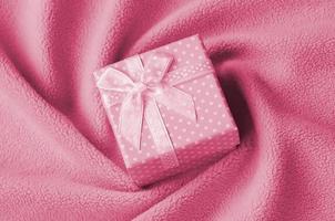een klein geschenk doos in roze met een klein boog leugens Aan een deken van zacht en harig licht roze fleece kleding stof met een veel van Verlichting vouwen. beeld afgezwakt in viva magenta, kleur van de jaar foto