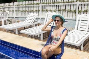 vrouw vervelend een een stuk zwempak en blauw Hoes omhoog hangende uit in de buurt een zwembad Aan een zonnig dag foto