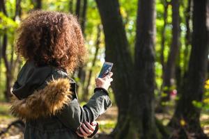 schattig gekruld haar- tiener meisje met smartphone in herfst Woud foto