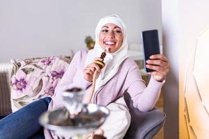 moslim vrouw roken shisha Bij huis en nemen een selfie met haar mobiel telefoon. moslim jong vrouw genieten van terwijl roken nargiel. Arabisch meisje roken hookah foto