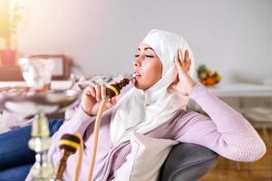 moslim vrouw roken shisha Bij huis. moslim jong vrouw genieten van terwijl roken nargiel. Arabisch meisje roken hookah foto