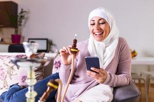 moslim vrouw roken shisha Bij huis en sms'en met haar vrienden. moslim jong vrouw genieten van terwijl roken nargiel. Arabisch meisje roken hookah foto