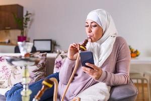 moslim vrouw roken shisha Bij huis en sms'en met haar vrienden. moslim jong vrouw genieten van terwijl roken nargiel. Arabisch meisje roken hookah foto