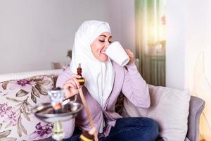 moslim vrouw roken shisha Bij huis en drinken koffie of thee. moslim jong vrouw genieten van terwijl roken nargiel. Arabisch meisje roken hookah foto