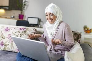 jong moslim vrouw hebben video telefoontje via laptop Bij huis. gelukkig glimlachen moslim vrouw zittend Aan bank, bankstel en gebruik makend van laptop Bij leven kamer Bij huis, aan het leren taal, video roeping. foto
