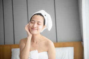 gelukkig jong Aziatisch vrouw toepassen gezicht lotions terwijl vervelend een handdoek en aanraken haar gezicht. dagelijks bedenken en huidsverzorging foto