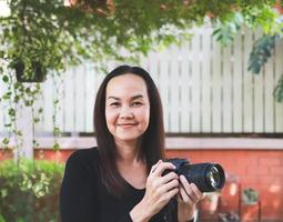 Aziatisch vrouw, vervelend lang mouw bovenkant, staand in de tuin, Holding dslr camera, glimlachen gelukkig. foto