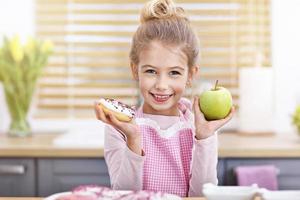 schattig weinig meisje kiezen tussen appel en donut in de keuken foto