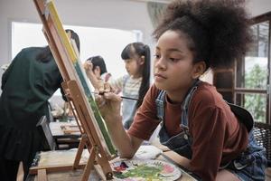 Afrikaanse Amerikaans meisje concentraten Aan acryl kleur afbeelding schilderij Aan canvas met studenten groep in kunst klas, creatief aan het leren met talenten en vaardigheden in elementair school- studio onderwijs. foto