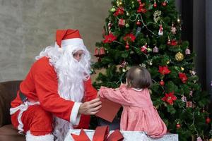 Kaukasisch baby is krijgen Cadeau van de kerstman claus Bij nacht door de ten volle versierd Kerstmis boom voor seizoen viering gebruik foto