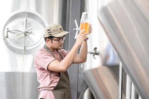 jong Aziatisch arbeider inspecteren brouwerij kwaliteit met een glas van ambacht bier evalueren zichtbaar uiterlijk na voorbereiding terwijl werken in een verwerken ambacht brouwerij. foto