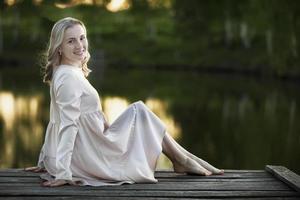 mooi jong blond vrouw Aan een houten brug in de buurt de rivier- poseren, op zoek Bij de camera. foto