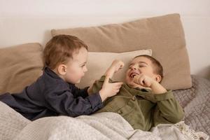 twee weinig en schattig Kaukasisch jongens spelen samen Aan de bed Bij huis. interieur en kleren in natuurlijk aarde kleuren. knus omgeving. kinderen hebben plezier, twee broers lastig elk ander. foto