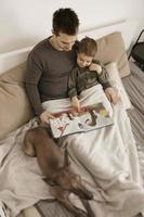 vader en zoon lezing een boek Aan de bed Bij huis. jong aantrekkelijk Mens en weinig jongen met hond resting in slaapkamer. natuurlijk aarde kleuren. knus omgeving. vader leest een fee verhaal voor zijn kind. foto