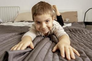 portret van klein, glimlachen Kaukasisch jongen Aan de bed Bij huis. schattig kind ontspannend, resting in slaapkamer. positief emoties. knus en modern interieur. natuurlijk, aarde kleuren. gewoontjes kleren. foto