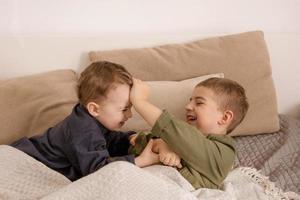 twee weinig en schattig Kaukasisch jongens spelen samen Aan de bed Bij huis. interieur en kleren in natuurlijk aarde kleuren. knus omgeving. kinderen hebben plezier, twee broers lastig elk ander. foto