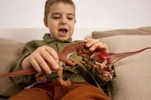 weinig en schattig Kaukasisch jongen spelen met dinosaurussen Bij huis. interieur en kleren in natuurlijk aarde kleuren. knus omgeving. kind hebben pret met speelgoed. foto