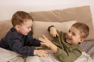 twee weinig en schattig Kaukasisch jongens spelen samen Aan de bed Bij huis. interieur en kleren in natuurlijk aarde kleuren. knus omgeving. kinderen hebben plezier, twee broers lastig elk ander. foto