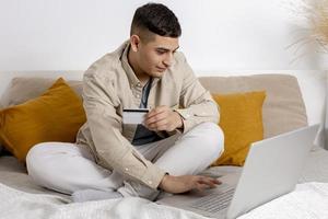 jong Mens met gewoontjes kleren surfing de web Aan zijn laptop computer en Holding credit kaart in zijn hand- proberen naar kopen iets online. gemakkelijk online boodschappen doen Bij huis, e-commerce. knus interieur. foto