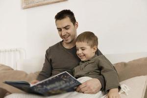 vader en zoon lezing een boek Aan de bed Bij huis. jong aantrekkelijk Mens en weinig jongen resting in slaapkamer. natuurlijk aarde kleuren. knus omgeving. vader leest een fee verhaal voor zijn kind. foto