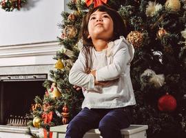 Aziatisch weinig meisje versieren de Kerstmis boom binnenshuis. de ochtend- voordat Kerstmis. portret liefhebbend meisje dichtbij omhoog. foto