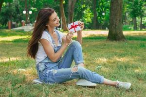 vrolijk jong meisje zit Aan een gras met geschenk in haar handen foto