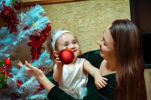 mooi weinig baby meisje met blauw ogen siert de Kerstmis boom met haar moeder foto