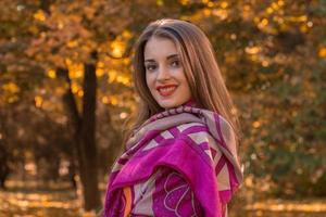 charmant jong meisje met rood lippenstift staat in de park sjaal Aan de schouders en glimlacht Bij kroepy plan foto