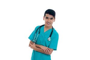 portret van jong vrolijk brunette Mens dokter in blauw uniform met stethoscoop glimlachen Aan camera gekruiste handen geïsoleerd Aan wit achtergrond foto