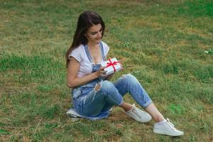 schattig brunette zit Aan de gras en houdt een klein geschenk verpakking foto