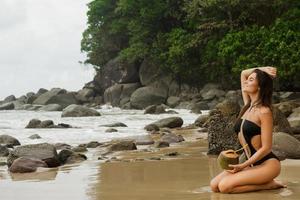 vrouw zittend Aan de rotsachtig strand met een kokosnoot cocktail foto