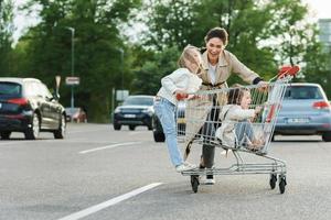 gelukkig moeder en haar dochters zijn hebben pret met een boodschappen doen kar Aan een parkeren veel naast een supermarkt. foto