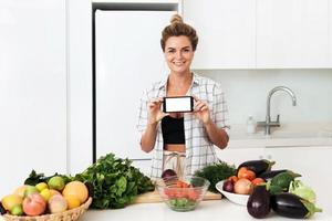 vrouw is Holding smartphone met blanco scherm voor uw ontwerp gedurende Koken in modern keuken foto