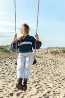 jong vrouw swinging Aan de schommel en op zoek weg Bij de zanderig strand. foto