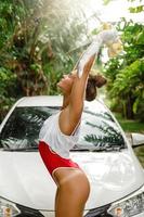 aantrekkelijk vrouw vervelend Bijsnijden top en rood bikini is het wassen haar wit auto in achtertuin foto