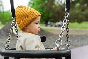 schattig baby jongen zittend Aan een schommel in openbaar park foto