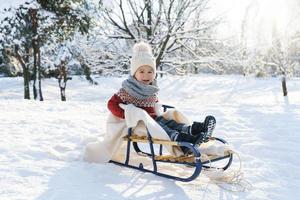 kleuter jongen zittend Aan de slee in een besneeuwd stad park gedurende zonnig winter dag foto