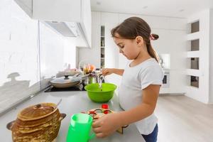 weinig schattig meisje is Koken Aan keuken. hebben pret terwijl maken cakes en koekjes. glimlachen en op zoek Bij camera. foto