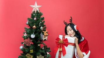 jong dame met rood santy kostuum Holding groot geschenk doos. de tafereel heeft een Kerstmis boom en een rood achtergrond. foto