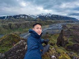 jong Aziatisch mannetje wandelaar zittend en nemen selfie Aan top met berg in IJslands hooglanden Aan somber dag in zomer foto