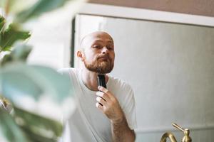 knap jong gebaard Mens trimmen zijn baard met machine in badkamer Bij huis foto