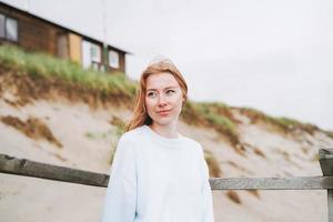 portret van jong rood haren vrouw in licht blauw trui Aan zand strand door zee in storm foto