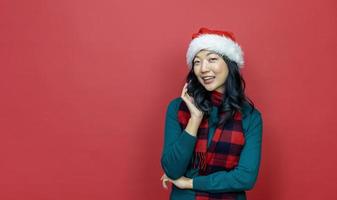 mooi glimlachen Aziatisch vrouw in warm Kerstmis trui en de kerstman hoed Aan rood achtergrond voor seizoen viering concept foto