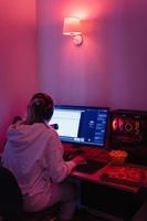 vrouw gamer of software ontwikkelaar zittend Bij de modern persoonlijk computer en aan het eten rommel voedsel Bij nacht in kamer met neon lichten foto