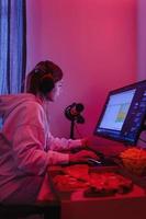 vrouw gamer of software ontwikkelaar zittend Bij de modern persoonlijk computer en aan het eten rommel voedsel Bij nacht in kamer met neon lichten foto